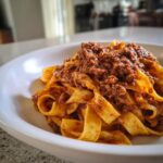 A close-up of fresh pappardelle pasta generously topped with rich, slow-cooked bolognese sauce in a white bowl.