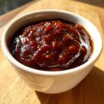 Close-up of thick, glossy, dark brown homemade bbq sauce with visible spices in a small white bowl.