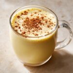 Close-up of creamy homemade eggnog topped with a dusting of cinnamon in a clear glass mug.