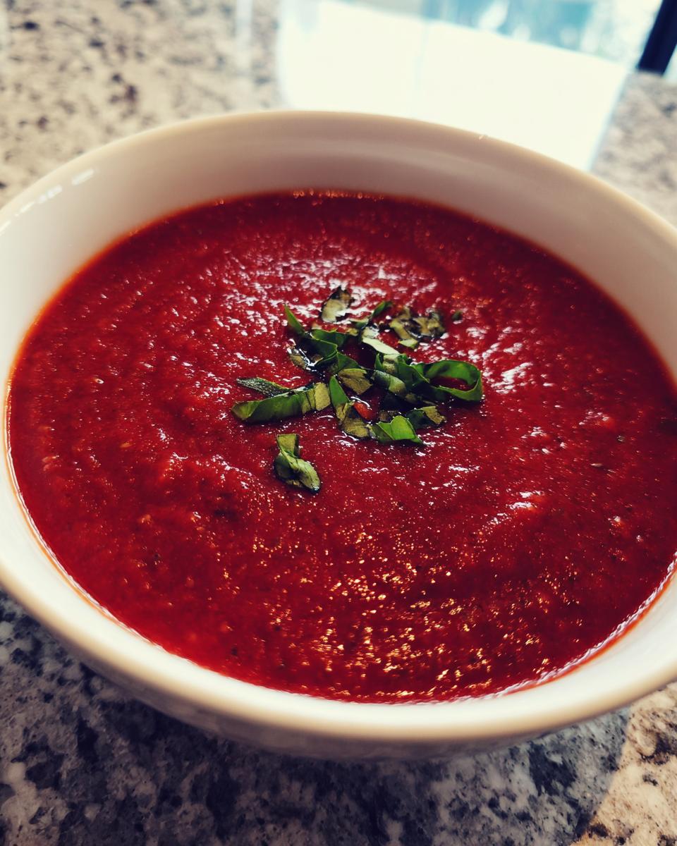Close-up of a bowl of vibrant, rich roasted tomato soup topped with fresh, chopped basil leaves.