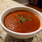 Close-up of a bowl of vibrant red roasted tomato soup, garnished with fresh chiffonade basil.