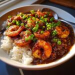 Close-up of a bowl of dark, rich shrimp etouffee served over fluffy white rice and topped with fresh green onions.