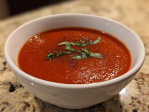 A close-up of a bowl of vibrant red roasted tomato soup, garnished with fresh basil and cracked black pepper.