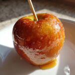 Close-up of a shiny, golden-brown caramel apples coated in crystallized sugar, resting on a white plate.