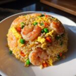 A mound of vibrant shrimp fried rice recipe featuring pink shrimp, scrambled egg, peas, and carrots on a white plate.