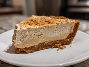 A close-up of a thick slice of creamy peanut butter pie with a graham cracker crust, topped with crumbs.