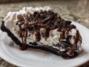 A decadent slice of ice cream pie featuring a dark crust, creamy filling with cookie pieces, topped with whipped cream, chocolate crumbles, and fudge sauce.