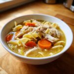 A close-up of a white bowl filled with rich broth, shredded chicken, egg noodles, and sliced carrots from the slow cooker chicken noodle soup.