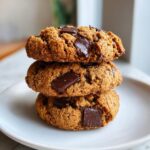 A stack of three thick, chewy vegan chocolate chip cookies loaded with melted chocolate chunks on a white plate.