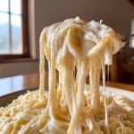 A fork lifts a generous portion of pasta heavily coated in thick, creamy olive garden alfredo sauce, showing long, gooey cheese pulls.