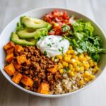 Close-up of a vibrant sweet potato taco bowl featuring seasoned ground meat, roasted sweet potatoes, rice, corn, avocado, and sour cream.