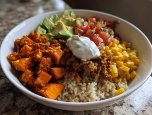 A vibrant sweet potato taco bowl featuring seasoned sweet potatoes, ground meat, brown rice, corn, avocado, salsa, and sour cream.