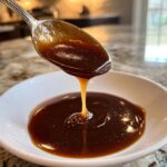 Close-up of thick, rich homemade brown gravy dripping from a silver spoon into a white bowl.