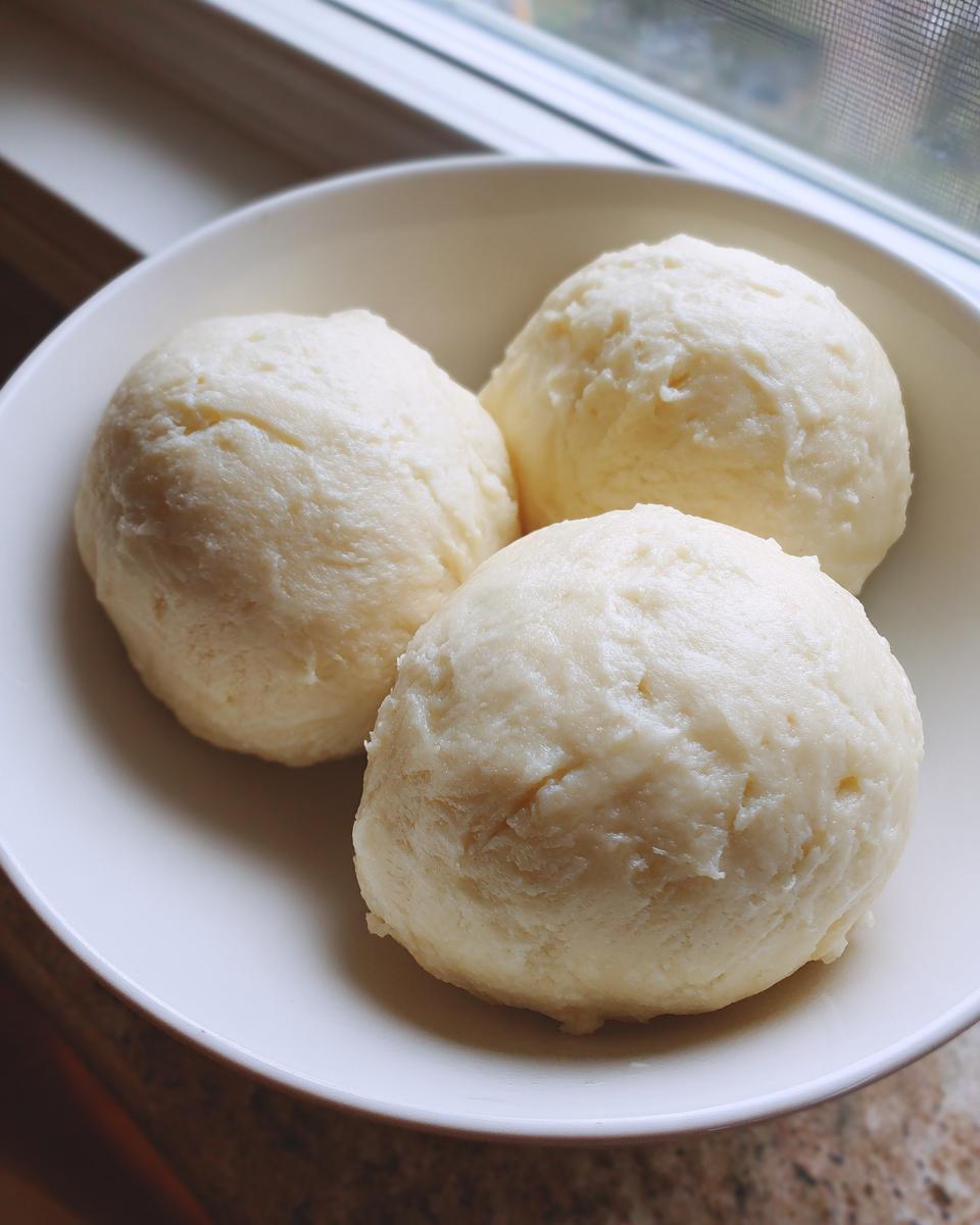 Three smooth, light-colored balls of freshly made fufu dough served in a white bowl near a window.