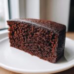 Close-up of a thick slice of ultra moist chocolate cake showing its dark, rich crumb texture on a white plate.