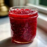 Close-up of a glass jar filled with bright red, textured freezer jam, catching sunlight.