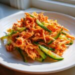 A close-up of a bright orange kani salad mixed with thin slices of fresh cucumber on a white plate.