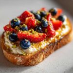 Close-up of a slice of ricotta toast topped with fresh strawberries, blueberries, honey, and chopped pistachios.