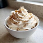 A white bowl filled with creamy, swirled banana ice cream topped with whipped cream and a sprinkle of spice.