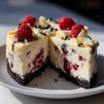 A cross-section view of a rich white chocolate raspberry cheesecake with a dark crust, topped with fresh raspberries.