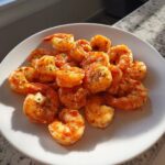 A mound of seasoned, cooked air fryer frozen shrimp piled high on a white plate, glistening in the sunlight.