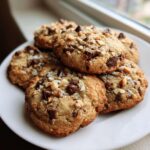 A stack of freshly baked almond joy cookies topped with chocolate chips, nuts, and coconut flakes, sitting on a white plate.