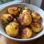 Close-up of golden brown, crispy roast potatoes seasoned with rosemary in a white bowl.