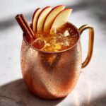 Close-up of a frosty copper mug filled with an apple cider moscow mule, garnished with apple slices and a cinnamon stick.