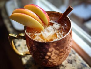 Close-up of a frosty copper mug filled with an apple cider moscow mule, garnished with apple slices and a cinnamon stick.