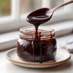 A spoonful of thick, glossy homemade chocolate syrup is dripping back into a glass jar, creating drips down the side.