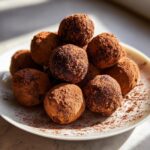 A close-up of several rich, dark bourbon balls generously dusted with cocoa powder, piled on a white plate.