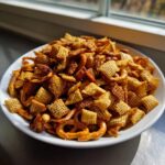 A white bowl overflowing with homemade, seasoned Chex party mix, featuring Chex cereal, pretzels, and nuts.