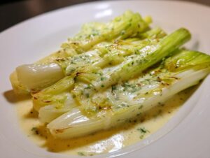 Close-up of tender braised leeks served on a white plate, covered in a creamy, herb-flecked sauce.
