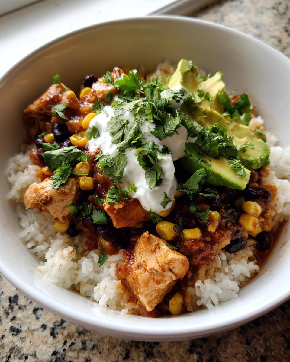 A close-up of a white bowl filled with white rice, topped with seasoned chicken, black beans, corn, sour cream, avocado slices, and fresh cilantro for a chicken burrito bowl.