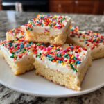 Close-up of several square christmas sugar cookie bars topped with thick white frosting and colorful rainbow sprinkles.