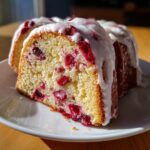 A close-up of a slice of moist cranberry orange cake, topped with a thick white glaze.