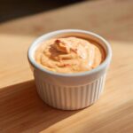 Close-up of creamy, orange-tinted burger sauce with red flecks, served in a small white ramekin on a wooden surface.