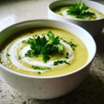 Close-up of a bowl of creamy, pale green celery soup, swirled with cream and topped with fresh parsley.