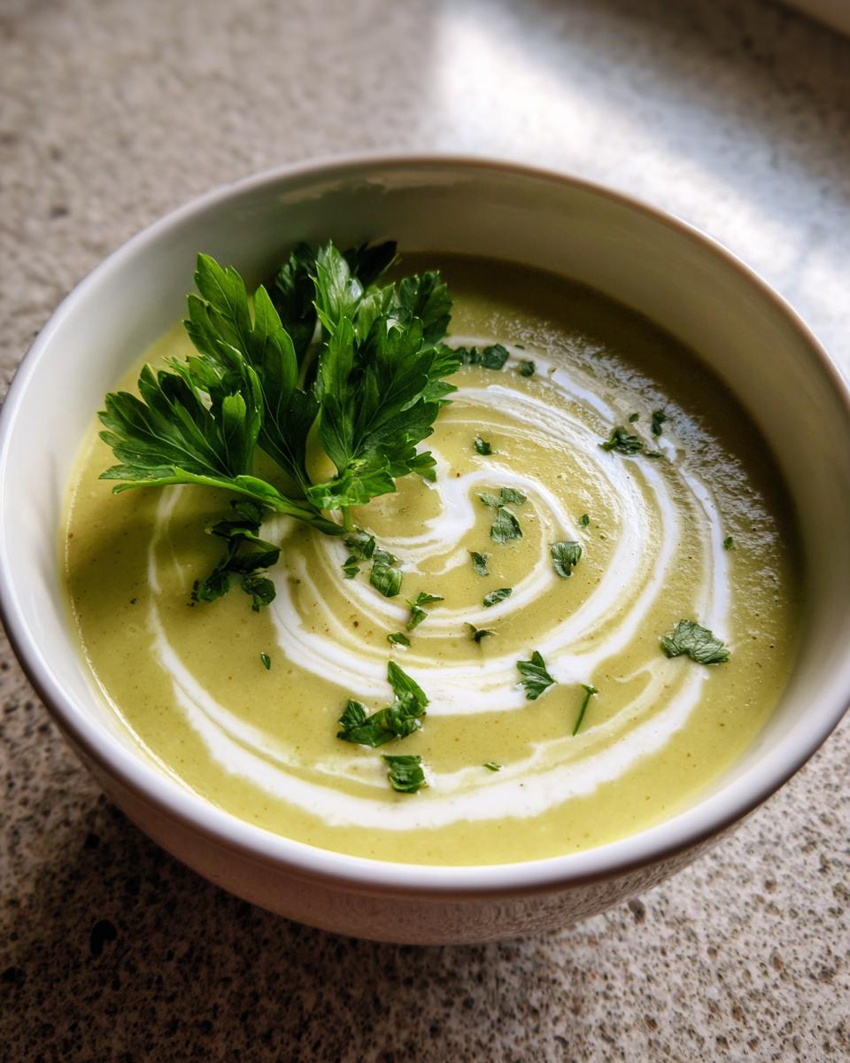 A close-up of creamy green celery soup topped with a white cream swirl and fresh parsley garnish.