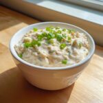 A white bowl filled with creamy clam dip, garnished generously with chopped green onions, sitting on a wooden surface.
