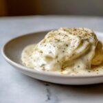 Close-up of a biscuit smothered in thick, creamy garlic sauce, seasoned with pepper and herbs.