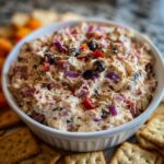 A white bowl filled with creamy hoagie dip mixed with diced meat, red onion, and black olives, surrounded by crackers.