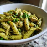Close-up of creamy pesto pasta featuring penne, chunks of chicken, and bright green peas served in a white bowl.