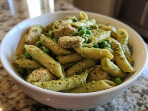 Close-up of creamy pesto pasta featuring penne, chunks of chicken, and bright green peas in a white bowl.