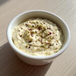 A close-up of creamy white pizza sauce topped with dried herbs and red pepper flakes in a small white ramekin.