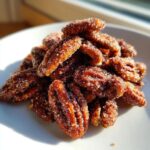 Close-up of a mound of glistening cinnamon sugar pecans piled high on a white plate, catching the sunlight.