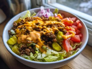A close-up of a delicious cheeseburger bowl featuring seasoned ground beef, melted cheese sauce, pickles, tomatoes, and onions over lettuce.