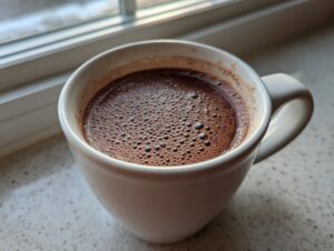 Close-up of rich, dark homemade hot cocoa with small bubbles on top, served in a white mug near a window.