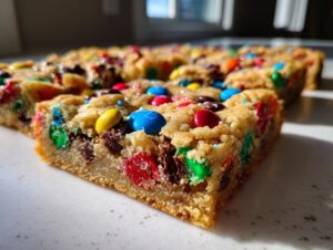 A close-up, low-angle shot of thick, chewy christmas cookie bars studded with colorful M&Ms and chocolate chips.