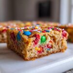 A close-up of a thick, chewy slice of Christmas cookie bars loaded with colorful M&M-style candies and sprinkles.